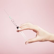 A hand holding a syringe set against a pink background.