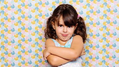 A little girl stands with her arms crossed and her tongue sticking out.