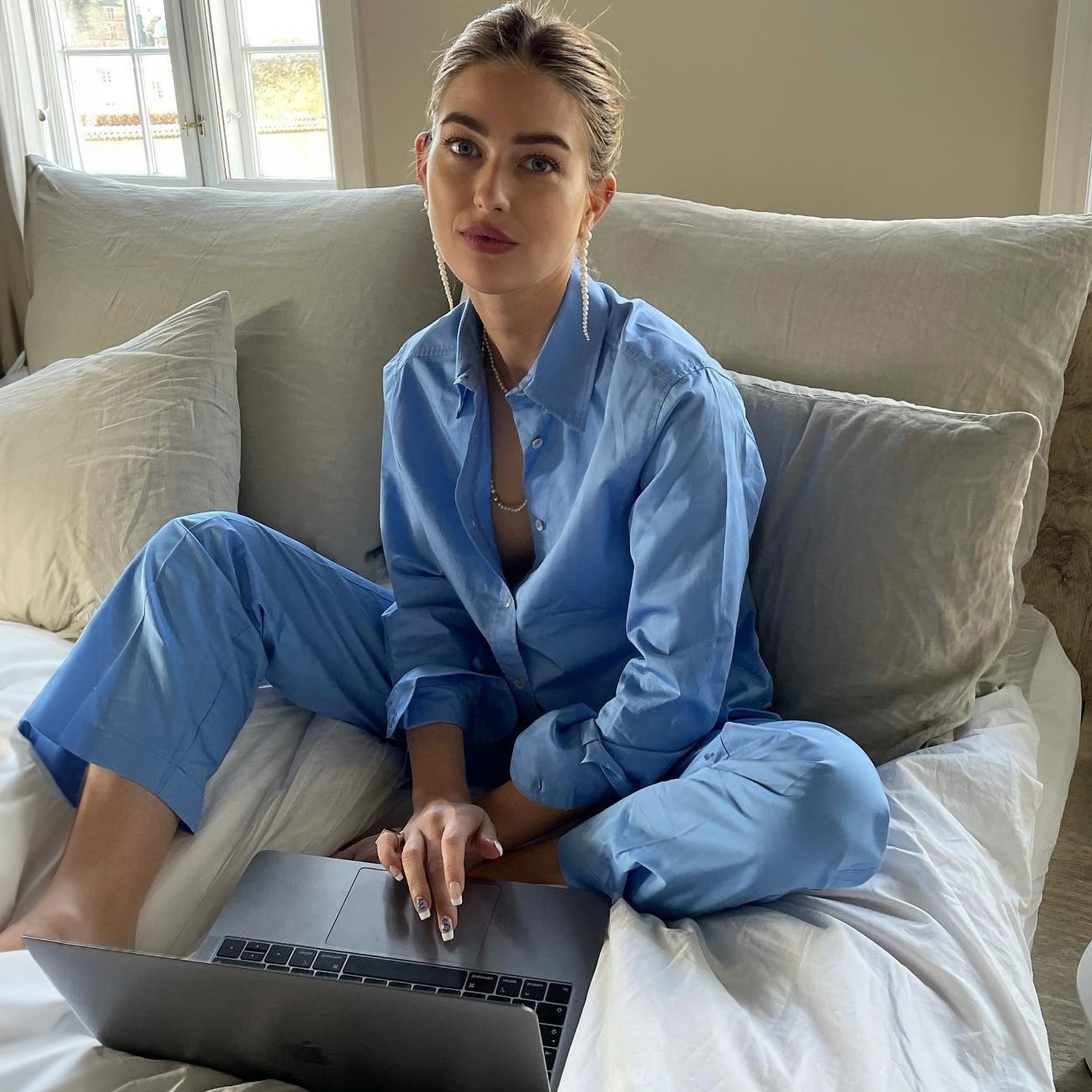 style influencer Emili Sindlev poses on a bed with neutral linen bedding wearing a light blue matching pajama set while scrolling on a laptop