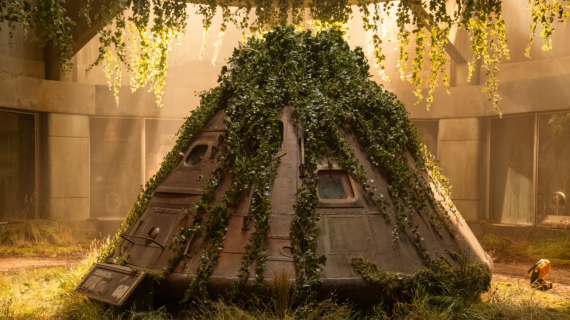 a space capsule overgrown by plants sits in the center of a sunlit rotunda