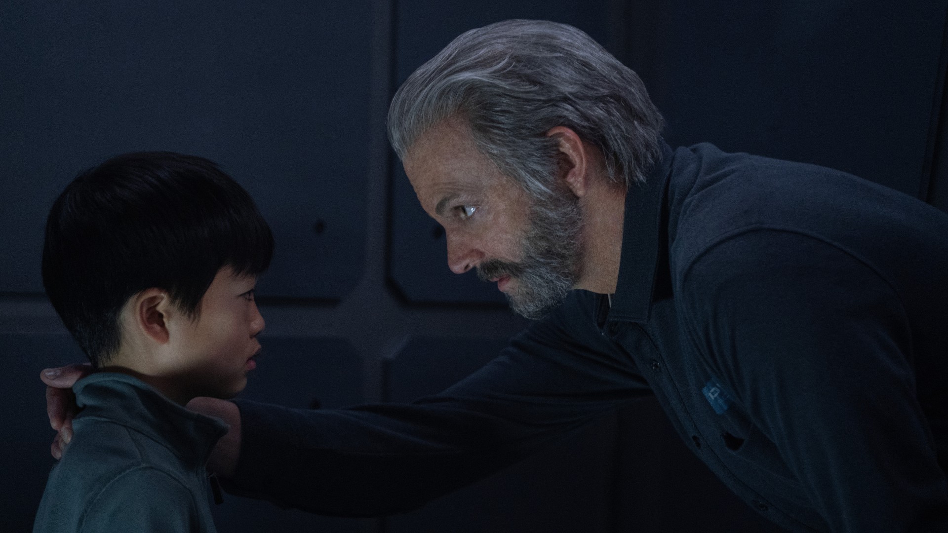 An older man (gray hair and beard) is crouching down and has his hand on the shoulder of a young boy in a caring manner.