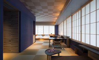 Interior view of a room at Hoshinoya, Tokyo, Japan featuring a checked ceiling and flooring, blue walls, grid style windows, two beds with white pillows and linen, a desk and a black and wooden table with matching chairs