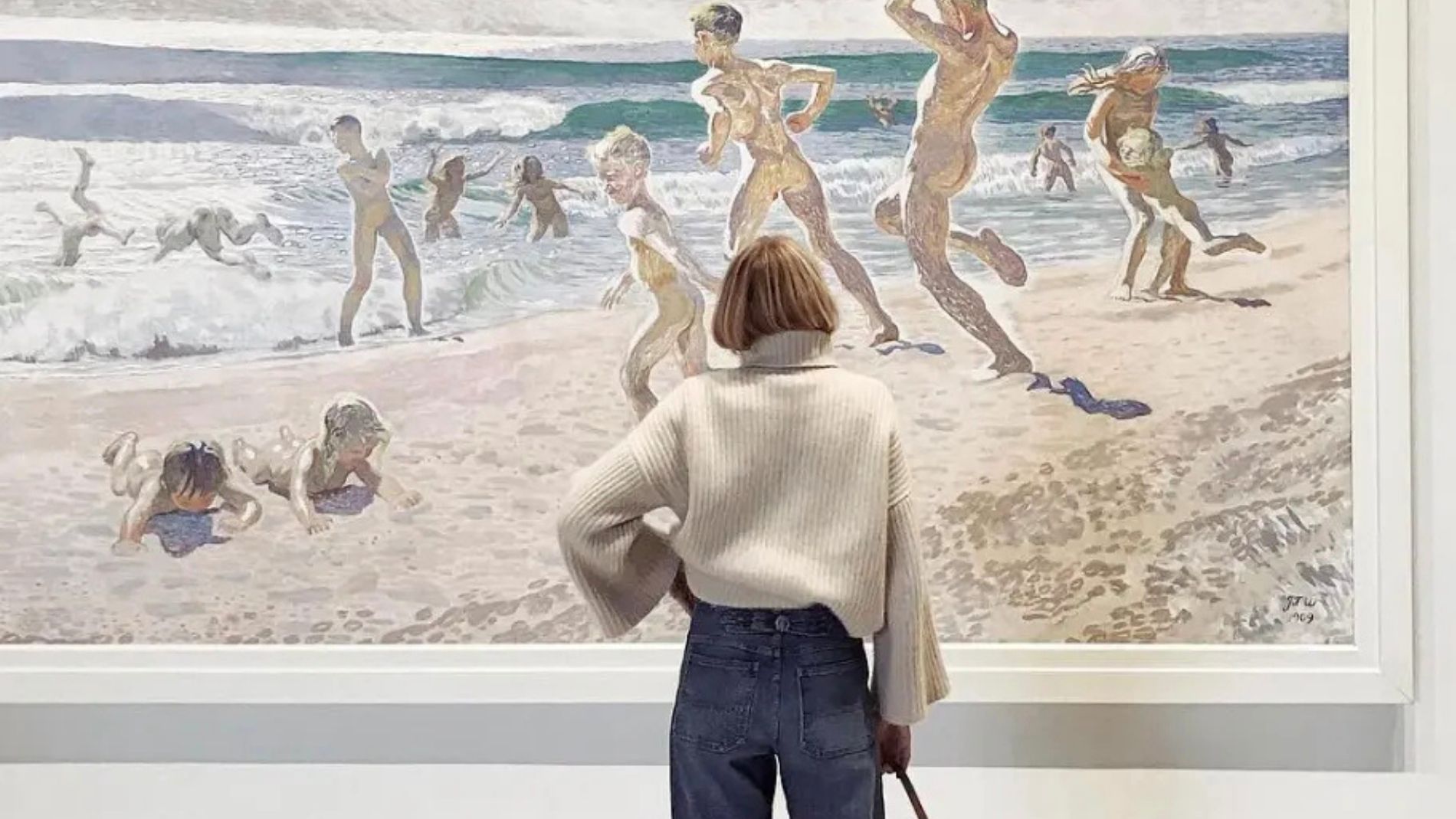 A woman looks at a painting while wearing a white sweater and jeans.