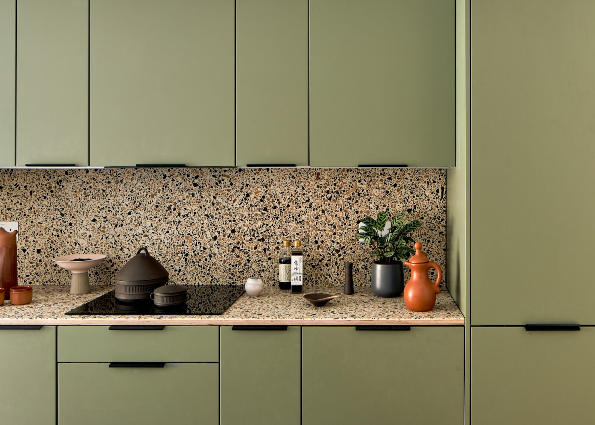A sage green colored kitchen with terrazzo worktops and backsplash. On the countertop is a stove, pots and other kitchen essentials.