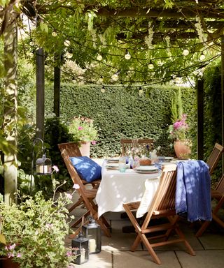 Wooden table and chairs in a small garden, surrounded by hedging and under a pergola with wisteria and string lights.