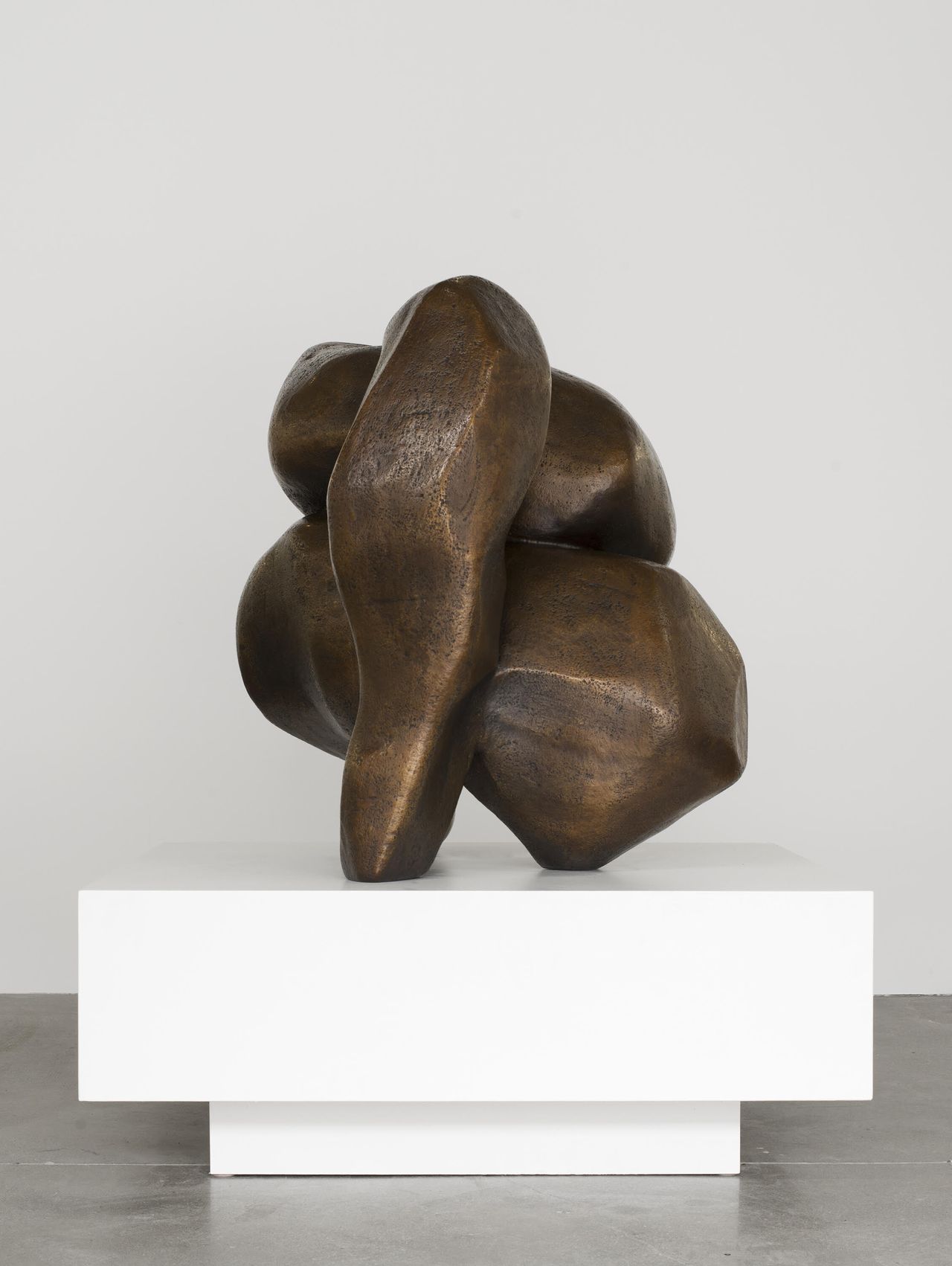 A bronze, abstract sculpture sits on a two-piece rectangular pedestal in a gallery.
