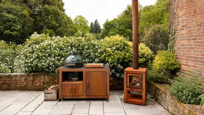A big green egg in an outdoor space with a wooden holder that has cabinets. There is also a fireplace on the side. There are lots of plants and flowers surrounding the grill 