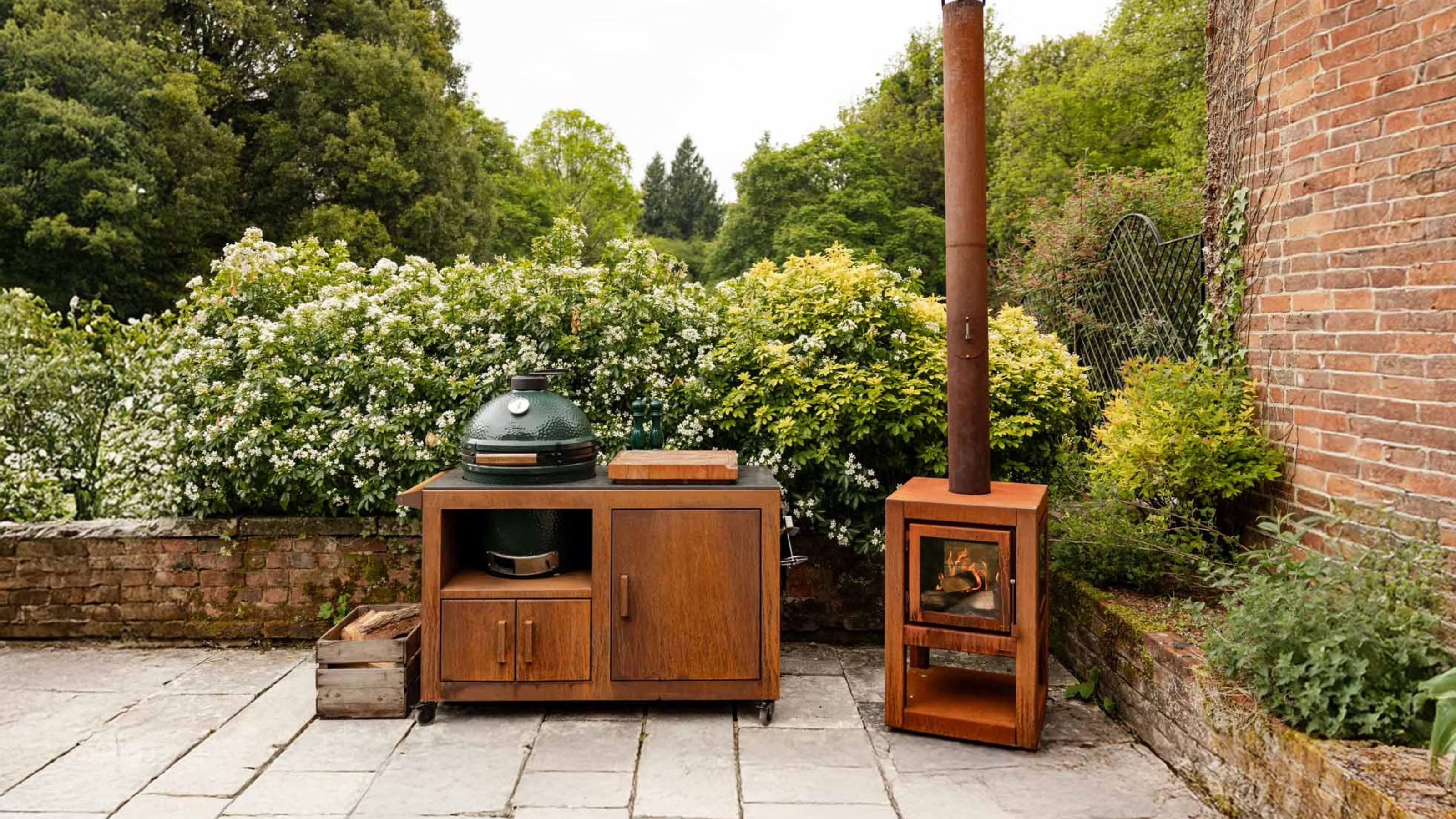 A big green egg in an outdoor space with a wooden holder that has cabinets. There is also a fireplace on the side. There are lots of plants and flowers surrounding the grill 
