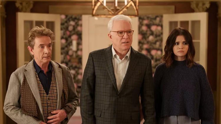 Scene from Only Murders in the Building: Three characters—two older men in blazers and a younger woman in a dark jumper—stand looking confused or alarmed in a warmly lit room with floral wallpaper.