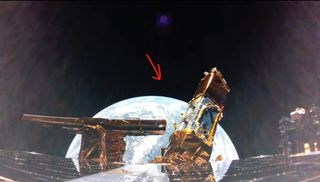 the dark, flat refelctive panels of a space craft line the foreground, with gold-foil wrapped copmonents sticking out in the middle. Behind, the blue round Earth in teh black of space. A red arrow above the planet points to a light pixel, which is the moon.