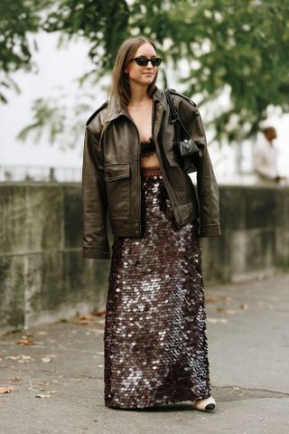 A woman at Paris Fashion Week Spring 2025 with her hair tucked into a leather jacket wearing a lace bra and sequin skirt