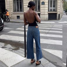 Sylvie wears cropped blue jeans, black kitten heels, and a black bodysuit.