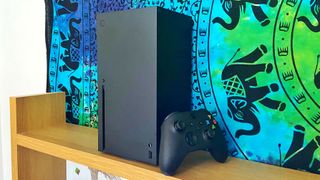 En Xbox Series X står på en trähylla med en tillhörande kontroller lutad mot ena sidan, framför en färgglad vägg.