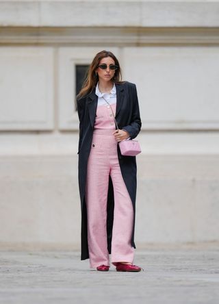 Gabriella Berdugo wears sunglasses, a black long coat, a white ruffled shirt, a pink striped jumpsuit with flared pants in tweed from Maje, red ballerina / ballet shoes from Jonak, a pink quilted leather vanity bag from Chanel, during a street style fashion photo session