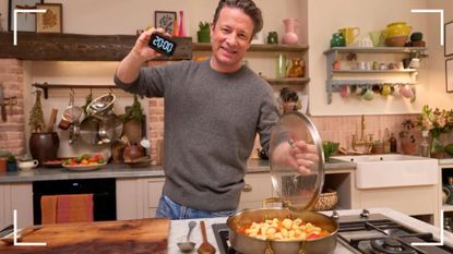 Jamie Oliver in his colourful kitchen cooking and holding up a timer