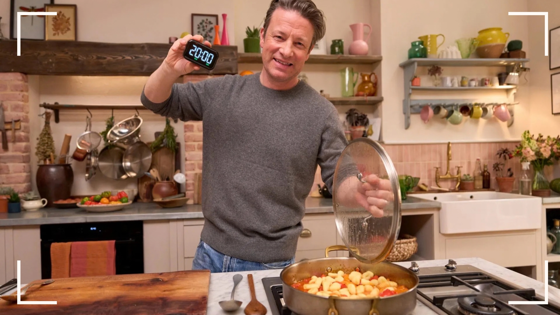 Jamie Oliver in his colourful kitchen cooking and holding up a timer