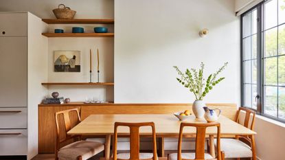 A white dining room with large windows, a wood table with wood chairs, a vase of stems, a bowl of fruit, floating wood shelves with bowls, art, candles, and a basket