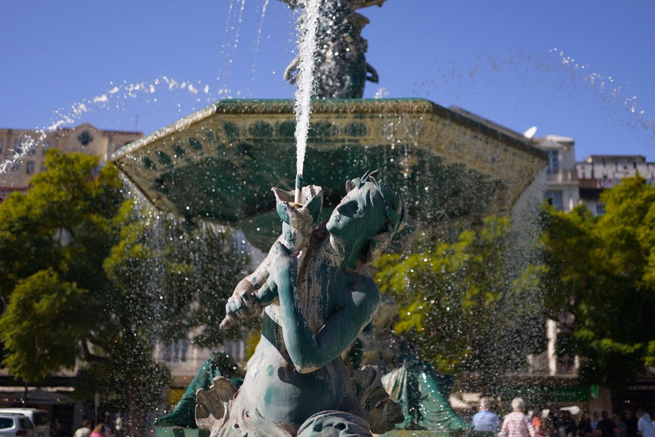 Decorative fountain figure spraying water, with droplets frozen in mid-air against trees and surrounding buildings.