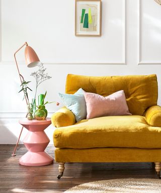 A mustard yellow large armchair with a pale pink and blue throw pillow, a wavy pink side table next to it with green glass vases, with a pink floor lamp next to it and a white wall with framed green wall art behind