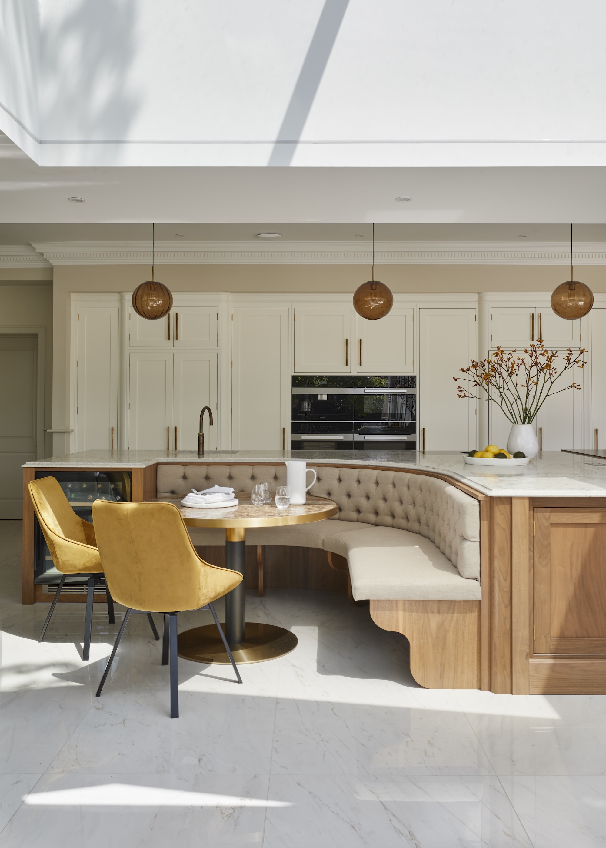 A cream kitchen with white marble flooring, a beige bench seat and round breakfast area, with a large sky light above.