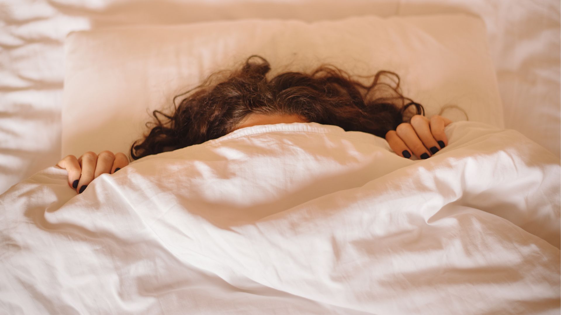 A woman lies on her back in bed, pulling a while duvet up over her face so it is obscured.