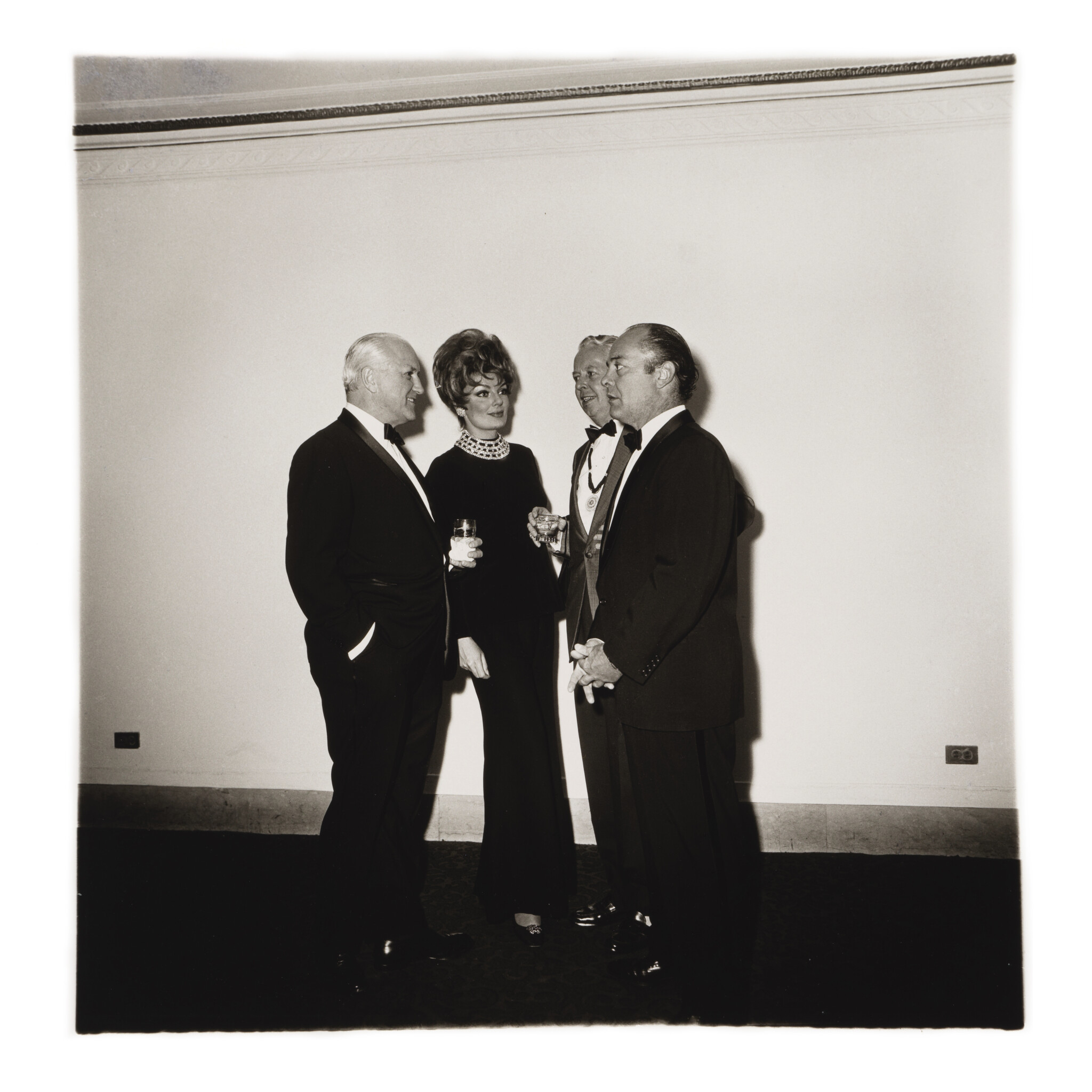 A black and white photograph captures four individuals in formal evening wear, including three men in tuxedos and a woman in a dark gown with a large pearl necklace, conversing at a social gathering.