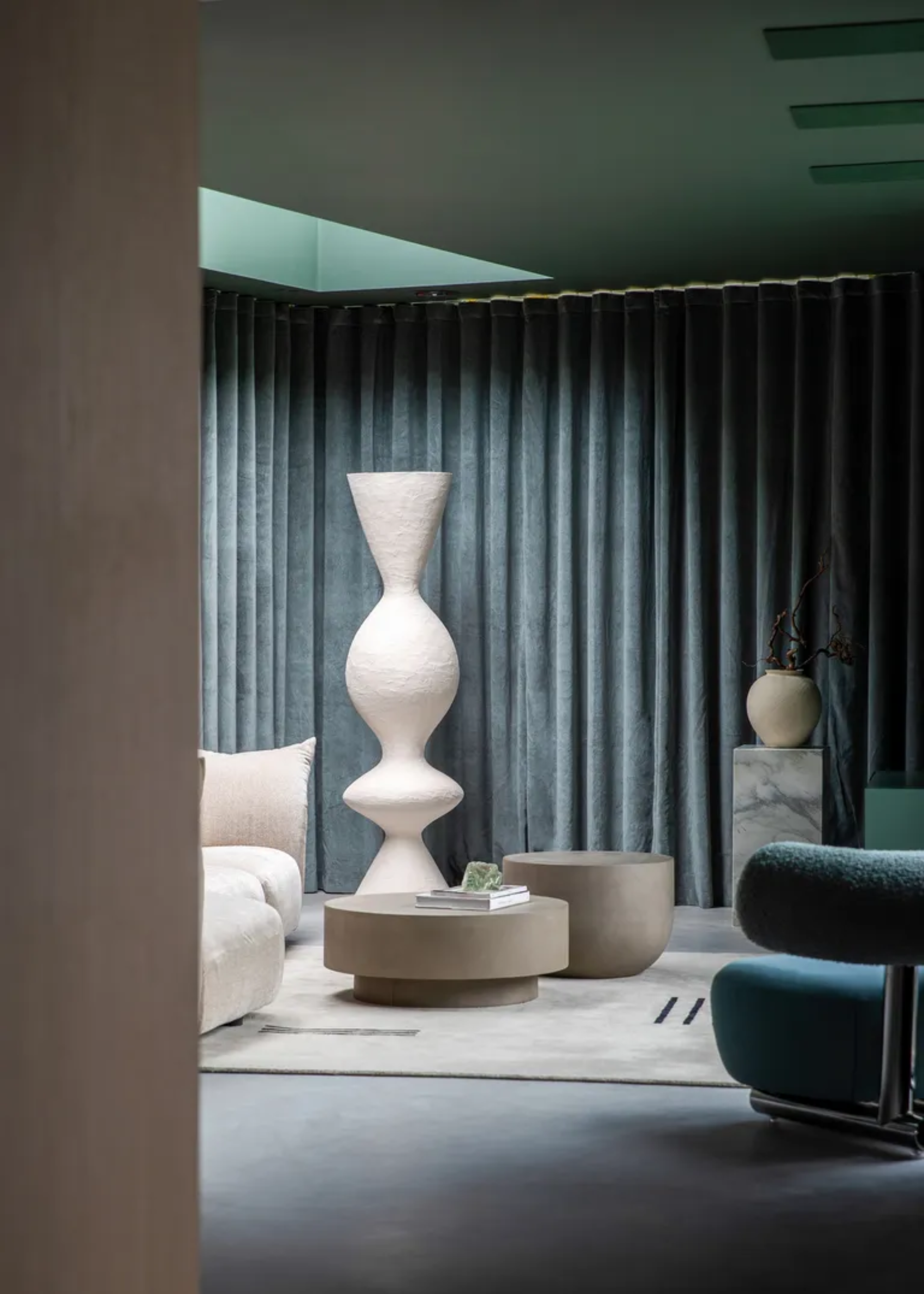 A living room with blue blackout curtains, blue floors, a blue fuzzy chair, a white sofa, a set of sandy coffee tables and a gray rug by a white structural decor piece
