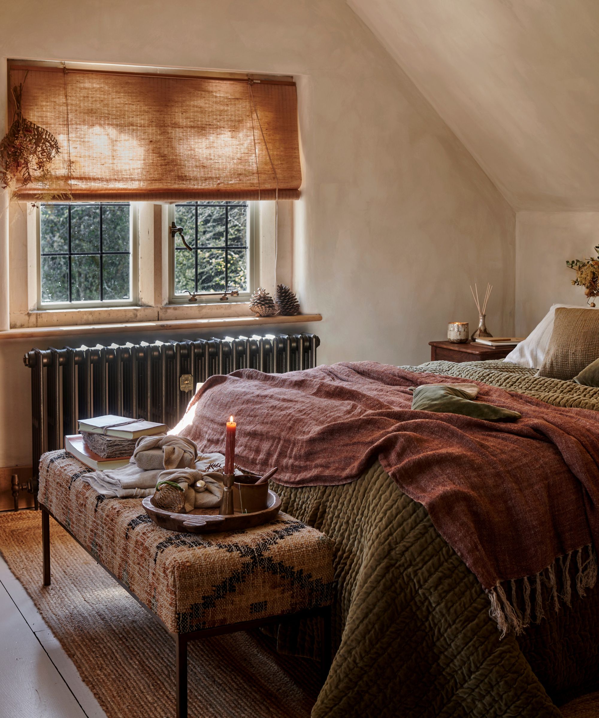 A cozy limewashed attic bedroom, with a cast iron radiator below a window. A bed with linen bedding in front