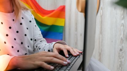 A woman types on a computer near a Pride flag