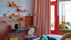 A colorful bedroom with baby blue and dusty rose walls, floating orange shelves with vases and trinkets, green wall lamps, a blue study with a black and white chair. A red door, a striped colorful curtain and a bay window with pillows.
