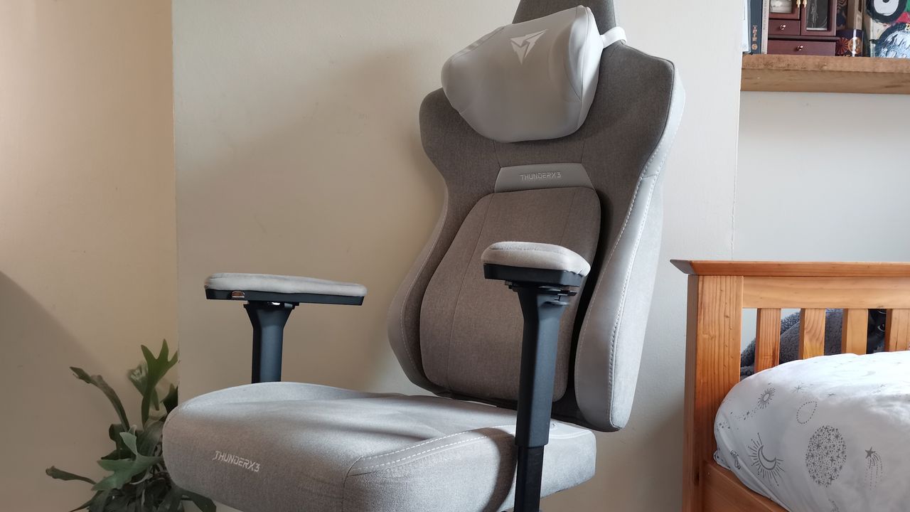 The ThundercoreX3 Core gaming chair in grey in front of a pale wall, next to a wooden bed frame
