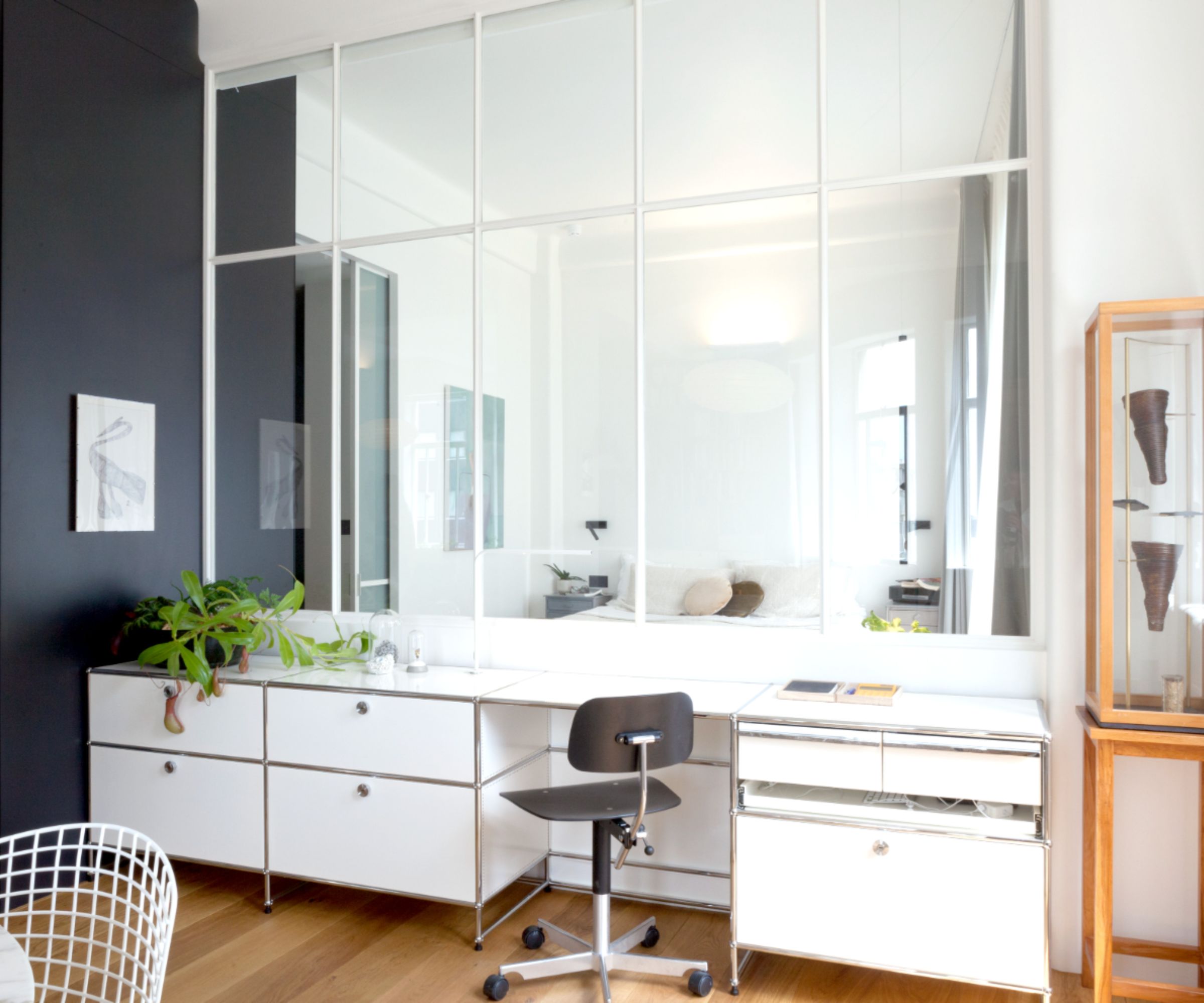 A sleek white desk with modular drawers and a black chair is set against a large internal window, bringing light into the compact home office nook.