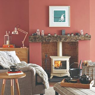 A terracotta-painted living room with a lit wood burning stove and a grey armchair covered with a knitted throw