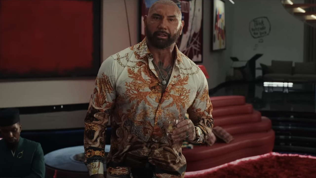 Dave Bautista berbicara dengan minuman di tangan, sementara Leslie Odom Jr. dalam Glass Onion a Knives Out menonton teka-teki tersebut.