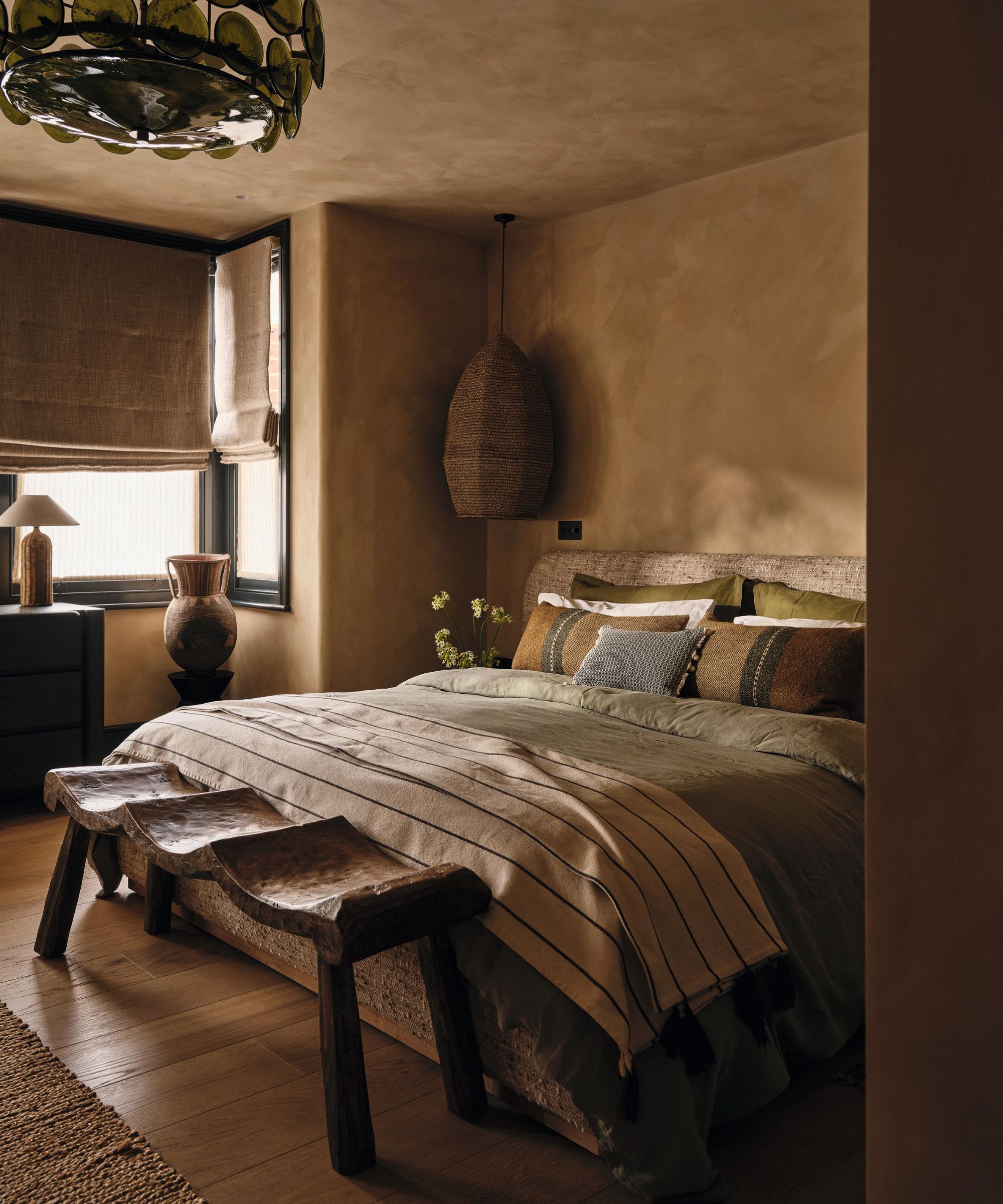 a dark beige limewashed bedroom with a linen bed, rustic wooden furniture, green glass chandelier and rattan pieces