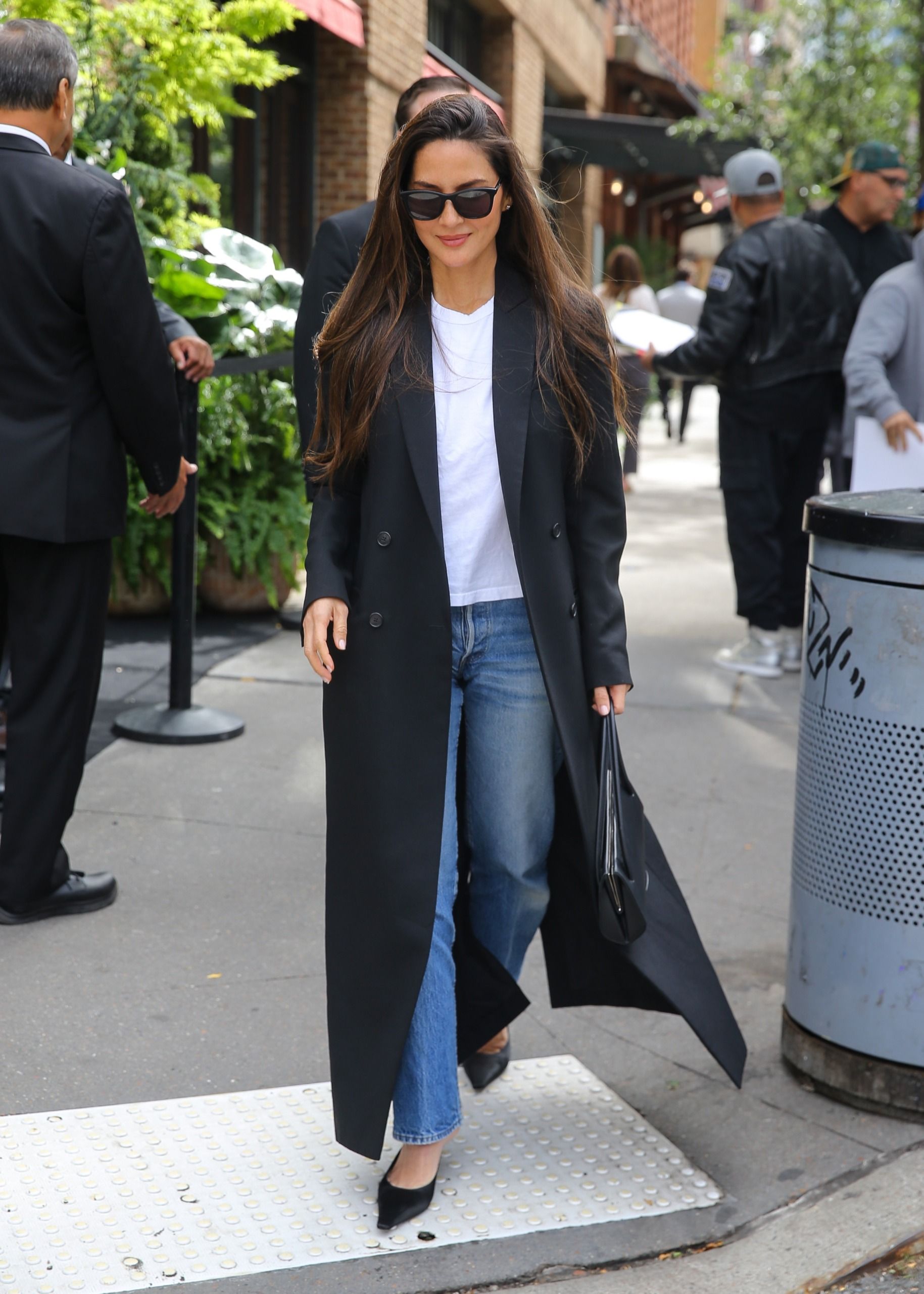 Olivia Munn wears a long black coat, white tee, jeans, and black satin heeled mules.