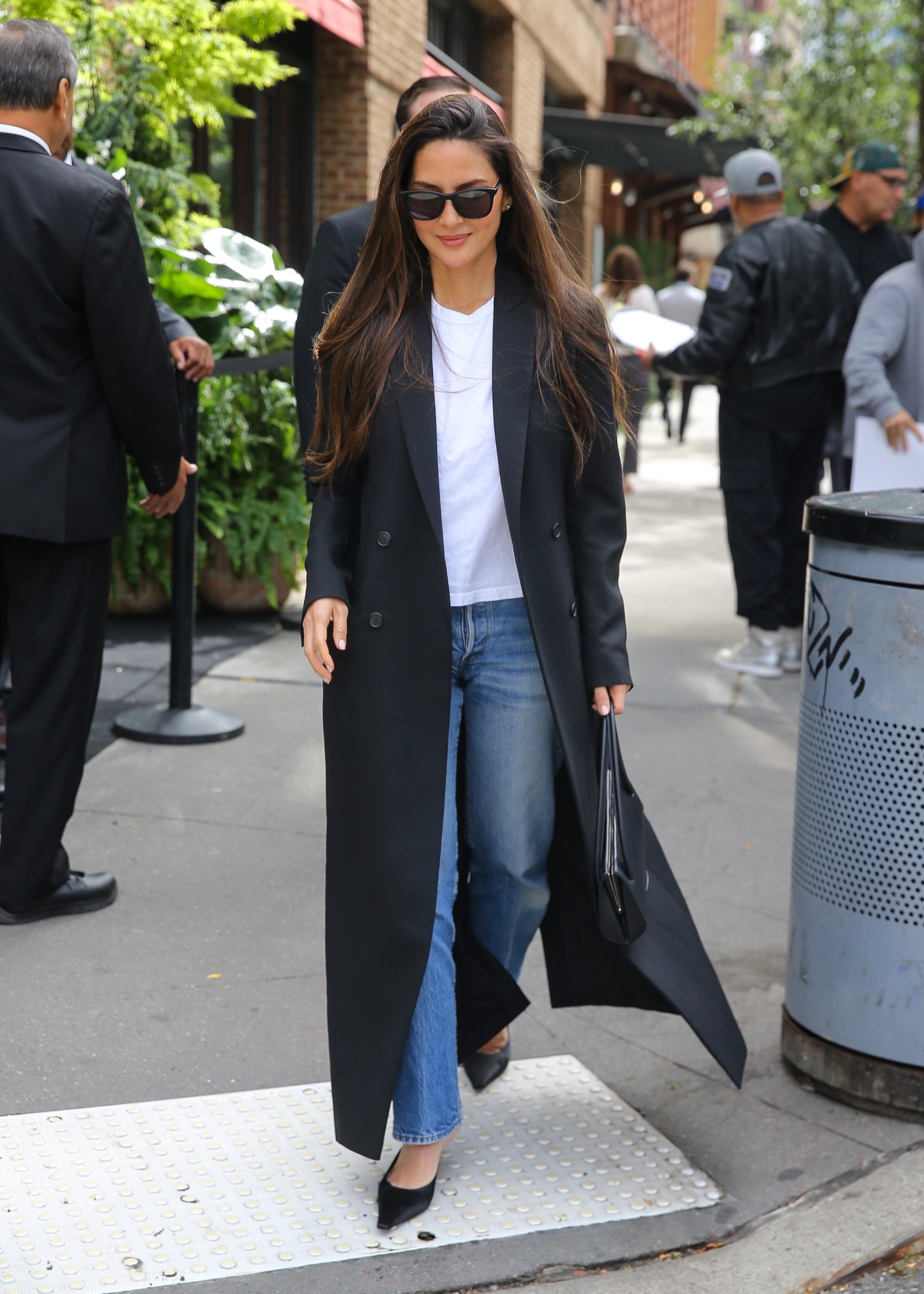 Olivia Munn wears a long black coat, white tee, jeans, and black satin heeled mules.