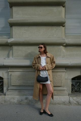 Spring trench coats shown in a photo of a woman standing in street wearing black sunglasses, a tan trench coat, white mini dress, a black shoulder bag, and black ballet flats