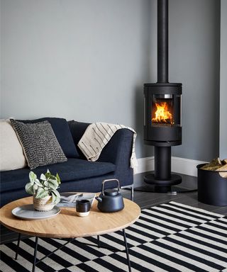 black corner log burner in room with vertical flue in room with black and white striped rug, blue sofa and birch table with black legs