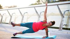 A woman performs a side plank with elevated leg exercise on a yoga mat outdoors. Her left forearm and foot rest on the ground, while the rest of her body is elevated. Her right hand points upwards and her right leg is elevated parallel to the floor. She is wearing a vest, leggings and sneakers. Behind her we see metal railings. 