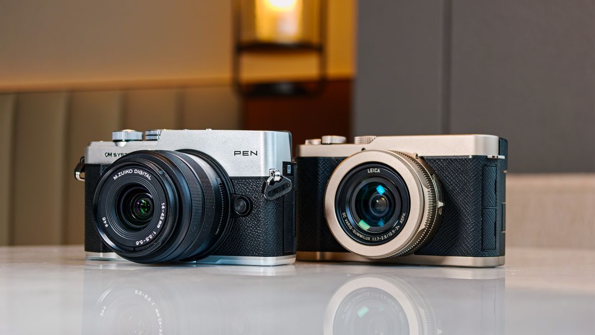 OM System PEN vs Panasonic Lumix L10: two micro four thirds cameras side by side on a white marble surface with lamp light in the background
