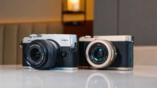 OM System PEN vs Panasonic Lumix L10: two micro four thirds cameras side by side on a white marble surface with lamp light in the background