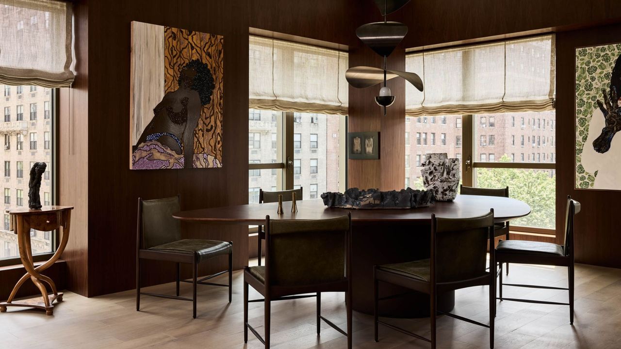 A brown dining room with large windows, Roman blinds, hanging paintings, a large dining table and a structured pendant