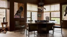 A brown dining room with large windows, Roman blinds, hanging paintings, a large dining table and a structured pendant