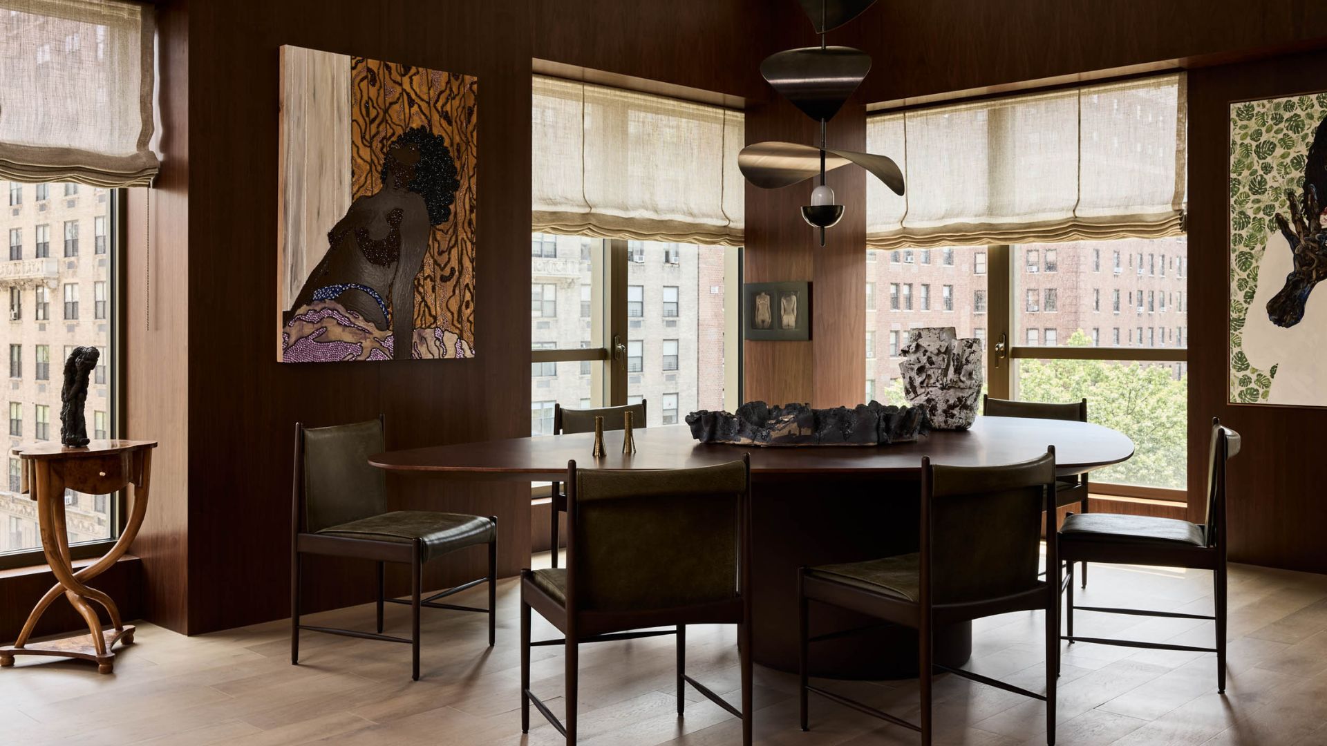 A brown dining room with large windows, Roman blinds, hanging paintings, a large dining table and a structured pendant