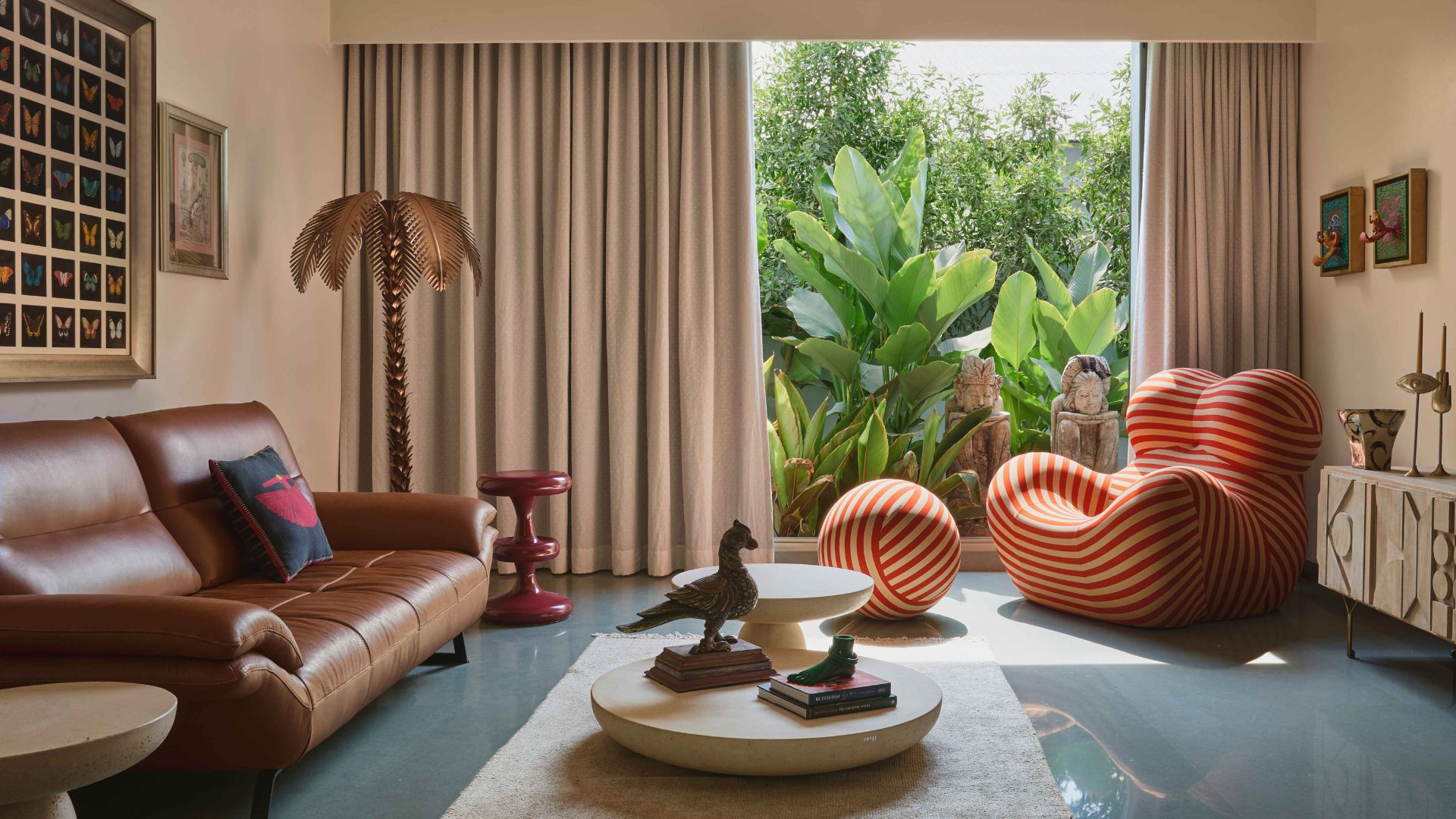 A bright living room with a glass window looking into the garden, a striped modular chair with a spherical leg rest, a low coffee table, a red side table, a leather couch, a sideboard, and a standing lamp 