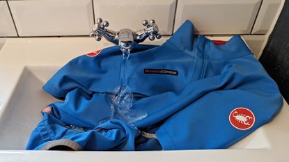 Blue Castelli Gabba Jacket in a sink with water pouring on top of it out of the tap 