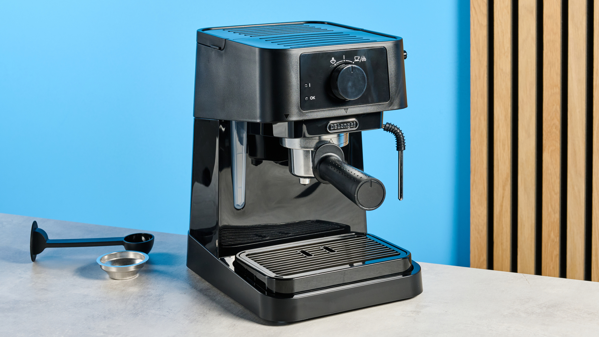 the de&#039;longhi stilosa espresso machine in black, with one portafilter, a dial for espresso and milk steaming, and a plastic tamper, photographed against a blue background