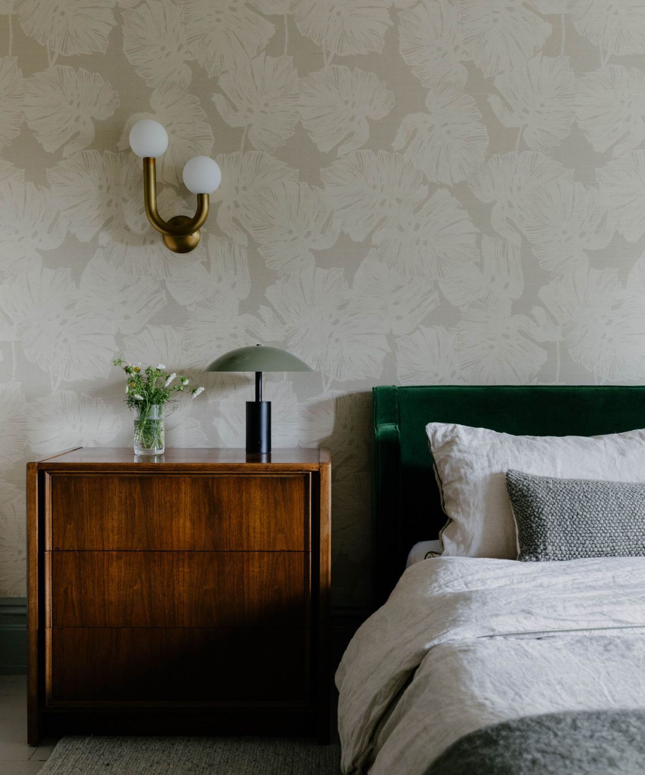 A bedroom with white and gray wallpaper, a green velvet headboard and a wooden side table