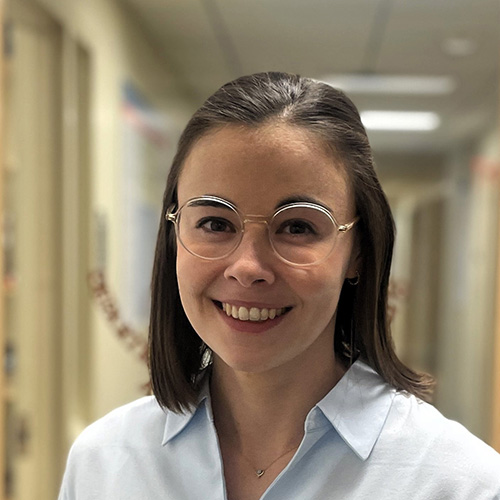 A woman with short brown hair and round glasses wearing a light blue button up shirt smiles for the camera.